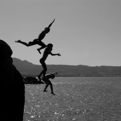 black-and-white photo of three people jumping off a cliff into the water